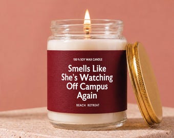 May include: A lit soy wax candle in a clear glass jar with a gold-colored metal lid. The label is deep red with white text that reads 100% SOY WAX CANDLE, Smells Like She’s Watching Off Campus Again, and BEACH RETREAT in a sans-serif font.