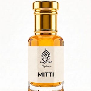 May include: A perfume bottle with a gold-coloured cap and a clear glass body. The bottle contains a light amber-coloured liquid. The label reads "AL UMAR Perfume" and "MITTI". The bottle has a unique shape.