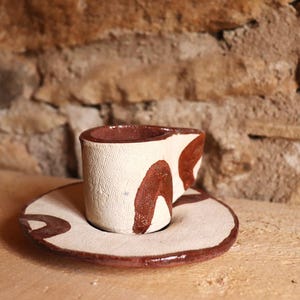 Peut inclure: Un ensemble tasse et soucoupe en céramique fait à la main. La tasse et la soucoupe sont blanc cassé avec des accents marron. La tasse est cylindrique avec un intérieur marron. La soucoupe est ronde avec un bord marron et un motif marron.