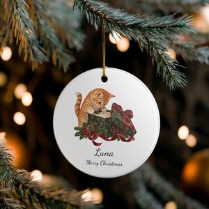 May include: A round white ceramic ornament featuring an illustration of an orange tabby kitten looking into a green gift box with a red ribbon. The text "Luna" and "Merry Christmas" is printed in black below the kitten. It hangs by a gold cord.