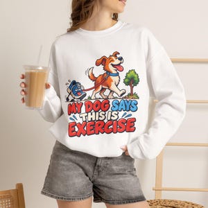 May include: A white long-sleeve sweatshirt featuring a graphic of a brown and white dog walking a blue sneaker on a leash. The text reads, "MY DOG SAYS THIS IS EXERCISE" in bold red and blue lettering. The design includes a small green tree.
