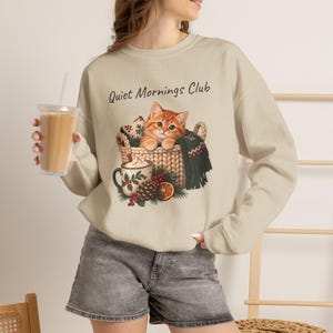May include: A beige crewneck sweatshirt featuring the text Quiet Mornings Club and a graphic of an orange tabby kitten sitting in a woven basket with a mug of hot cocoa, a pinecone, holly berries, and a slice of dried orange.