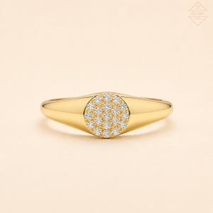 Op de afbeelding: Een gouden zegelring met een cirkelvormig vlak ingelegd met een cluster van kleine, heldere, rondgeslepen stenen. De ringband is glad en gepolijst, loopt taps toe naar de achterkant, met de merknaam ELARA in de rechterbovenhoek gedrukt.