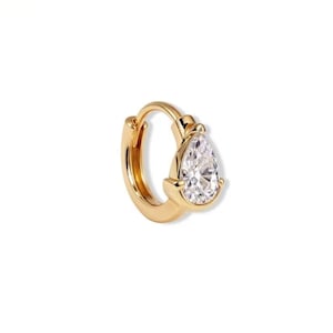 May include: A gold hoop earring featuring a single pear-shaped clear gemstone set in a prong setting. The hoop has a polished metallic finish and a hinged closure mechanism, showcasing a reflective surface and a faceted stone that catches light.