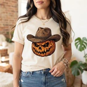 May include: A cream-coloured short-sleeve t-shirt featuring a graphic of a carved orange pumpkin wearing a brown cowboy hat. The pumpkin has a jagged, sinister facial expression with glowing eyes and a wide, toothy grin, centred on the chest.
