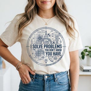 May include: A cream-colored short-sleeve t-shirt featuring a dark blue circular graphic design. The print includes technical engineering drawings of gears, a compass, and a caliper, with the text "I SOLVE PROBLEMS YOU DON'T KNOW YOU HAVE."