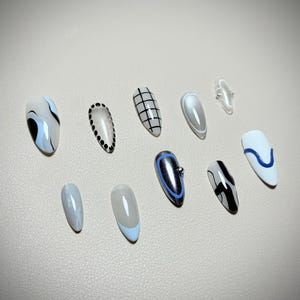 May include: A collection of ten artificial nails in various designs. The nails feature a range of colors, including white, black, blue, and silver. Designs include abstract swirls, grid patterns, and metallic accents. The nails are oval-shaped.