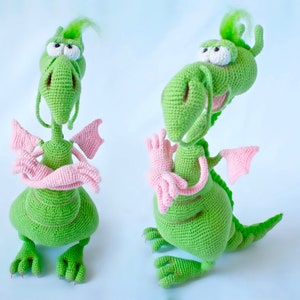 Puede incluir: Dos peluches de dragón verde hechos a crochet con alas rosas y un cuello largo verde. Los dragones tienen ojos blancos y están de pie sobre un fondo blanco.