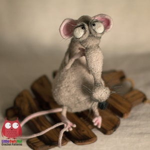 Pode incluir: Um rato cinzento de croch&ecirc; com olhos grandes e salientes e um nariz longo e fino. O rato est&aacute; sentado em uma plataforma de madeira.
