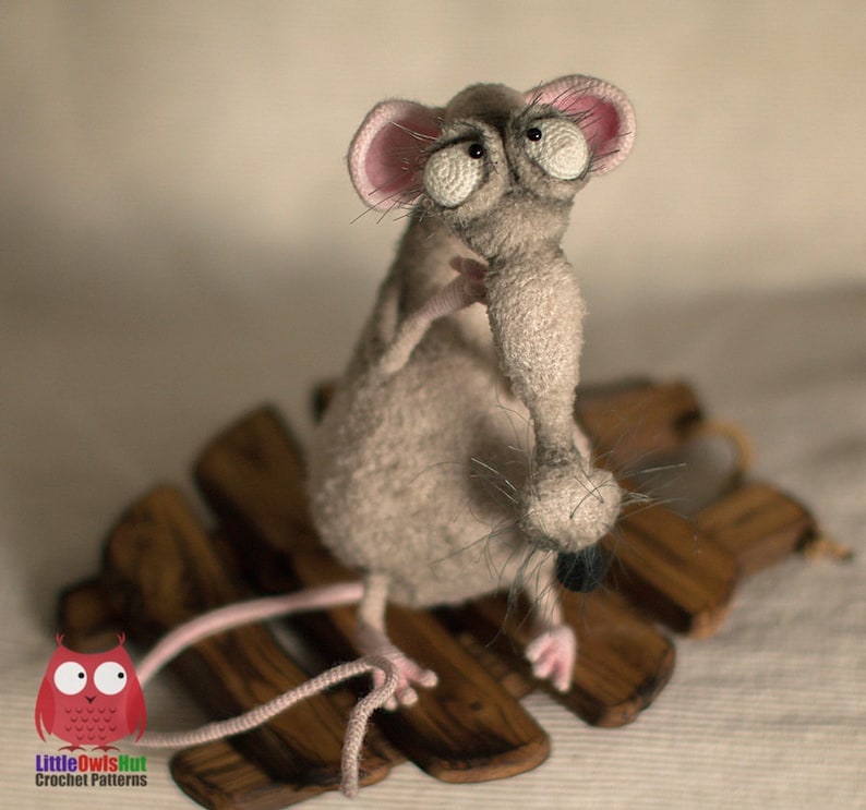 Pode incluir: Um rato cinzento de croch&ecirc; com olhos grandes e salientes e um nariz longo e fino. O rato est&aacute; sentado em uma plataforma de madeira.