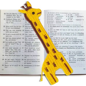 Può includere: Un segnalibro di giraffa all'uncinetto giallo con occhi marroni e un collo all'uncinetto marrone. Il segnalibro è posizionato su un libro aperto con testo in russo e inglese.