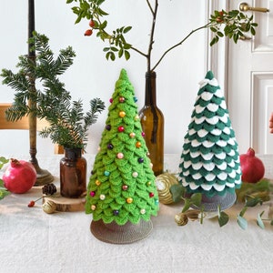 May include: Two crocheted Christmas trees, one green with colorful beads and one green and white, on a white table with candles and greenery.