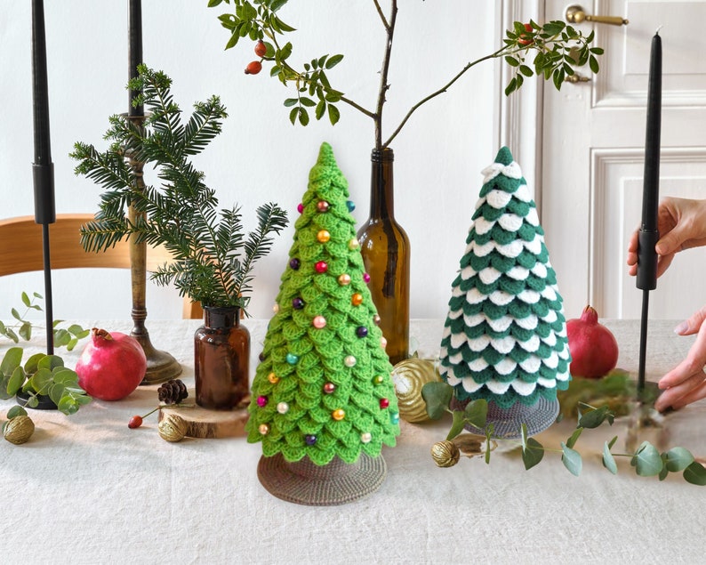 May include: Two crocheted Christmas trees, one green with colorful beads and one green and white, on a white table with candles and greenery.