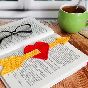 Pode incluir: Um marcador de página de crochê em forma de flecha com um coração vermelho na ponta. O marcador está sobre um livro aberto com uma chávena de chá e óculos na mesa.