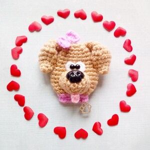 May include: A crocheted brown dog with pink accents and a bow, surrounded by red hearts.