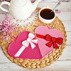 Könnte beinhalten: Zwei herzförmige Topflappen aus Häkelarbeit in Rosa, verziert mit Schleifen. Einer hat eine weiße Schleife und ein weißes Band, der andere eine rote Schleife und ein rotes Band. Eine weiße Teekanne, eine Tasse Tee und rosa Blumen sind ebenfalls vorhanden.