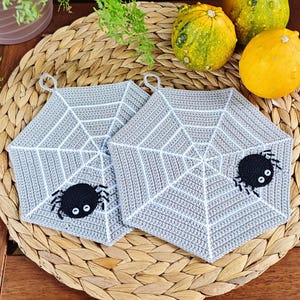 May include: Two gray crocheted pot holders shaped like spiderwebs with black spiders in the center. The pot holders are on a woven wicker surface.