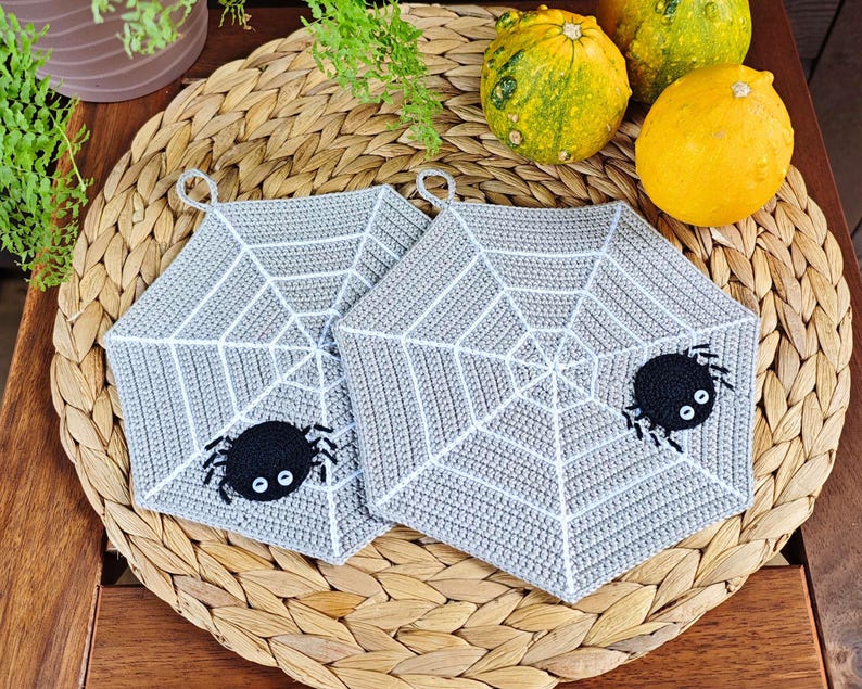 May include: Two gray crocheted pot holders shaped like spiderwebs with black spiders in the center. The pot holders are on a woven wicker surface.