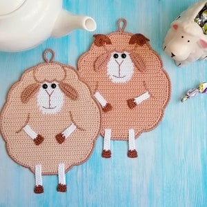 以下が含まれることがあります： 茶色と白の毛糸で編まれた羊の形をした鍋つかみが2つ。羊は黒い目と茶色の鼻をしています。