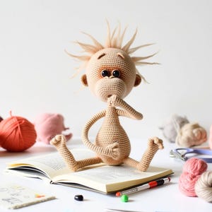 May include: A crocheted plush toy with brown yarn and large eyes. The toy is sitting on a book with a pencil and other yarn balls nearby.