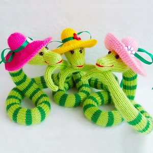 May include: Three knitted green snakes with striped patterns are wearing colorful hats. One snake has a pink hat, another has a yellow hat, and the third has a pink hat with a white flower.