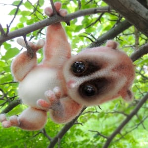 May include: A crocheted plush toy resembling a slow loris, hanging from a tree branch. The toy has large, black eyes, a white belly, and a pink and brown body.