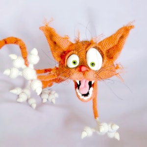Puede incluir: Un juguete de gato naranja de crochet con patas blancas y una boca muy abierta. El gato tiene ojos verdes y una lengua rosa.