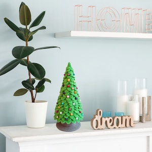 May include: A green crocheted Christmas tree with colorful ornaments sits on a white shelf with a wooden sign that says "dream". A potted plant with large green leaves is on the left side of the shelf.