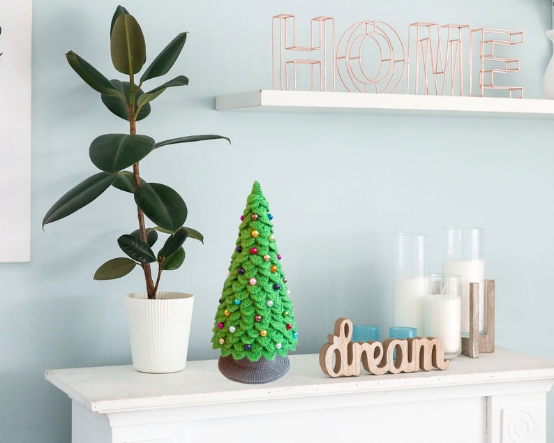 May include: A green crocheted Christmas tree with colorful ornaments sits on a white shelf with a wooden sign that says "dream". A potted plant with large green leaves is on the left side of the shelf.