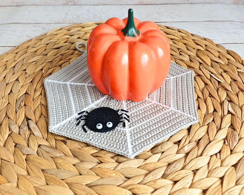 May include: A crocheted gray and white spiderweb pot holder with a black spider in the center. The pot holder is on a woven brown wicker surface with an orange pumpkin on top.