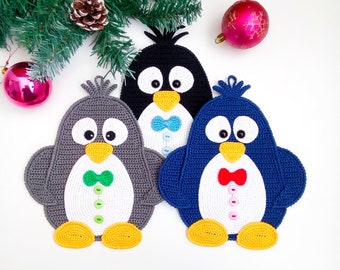 Könnte beinhalten: Drei gehäkelte Pinguin-Ornamente in Grau, Schwarz und Blau. Jeder Pinguin hat einen weißen Bauch, gelbe Füße, einen gelben Schnabel und schwarze Augen. Sie sind mit kleinen bunten Fliegen und Knöpfen in Grün, Hellblau und Rot verziert.