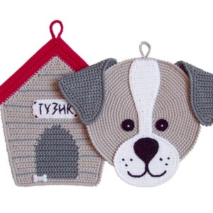 Puede incluir: Dos agarraderas de olla de ganchillo, una con forma de cara de perro y la otra con forma de caseta. La caseta tiene un techo rojo y las palabras "TUZIK" en la parte delantera. Ambas agarraderas son grises con detalles blancos.