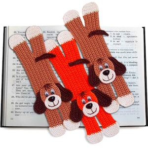 May include: Two crocheted dog-shaped bookmarks. One is brown with white accents and the other is orange with white accents. Both bookmarks are lying on an open book.
