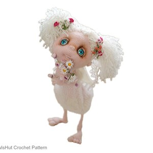 Puede incluir: Una mu&ntilde;eca de crochet blanca con cabello largo blanco, ojos azules y mejillas rosadas. La mu&ntilde;eca sostiene un ramo de flores blancas y lleva una corona de flores rosas.