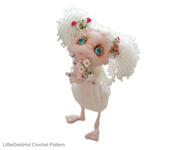 Puede incluir: Una mu&ntilde;eca de crochet blanca con cabello largo blanco, ojos azules y mejillas rosadas. La mu&ntilde;eca sostiene un ramo de flores blancas y lleva una corona de flores rosas.