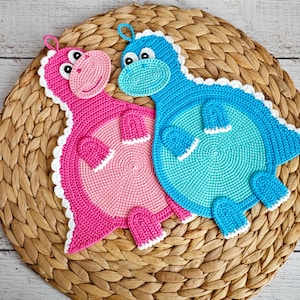 May include: Two crocheted dinosaur-shaped pot holders, one pink and one blue, with white trim. The pot holders are on a woven wicker surface.