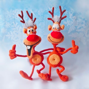 May include: Two crocheted reindeer toys with red noses and white fur on their heads. They are both smiling and giving a thumbs up.