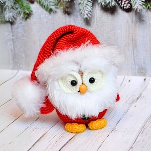 May include: A crocheted owl wearing a red Santa hat with white fur trim. The owl has white fur and black eyes. The owl is sitting on a white wooden surface.