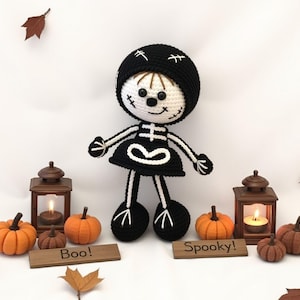 May include: A crocheted skeleton doll wearing a black dress and a black hat. The doll is standing on a white surface with two wooden lanterns and pumpkins around it. The lanterns have candles inside. The doll has a friendly face with black eyes and a stitched smile. The doll is holding its hands up in a welcoming gesture. The text "Boo!" and "Spooky!" is written on wooden signs in front of the doll.