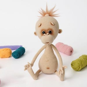 May include: A crocheted orangutan toy with brown eyes and a light brown body. The toy is sitting with its legs crossed and its arms outstretched. The toy is surrounded by balls of yarn in various colors.