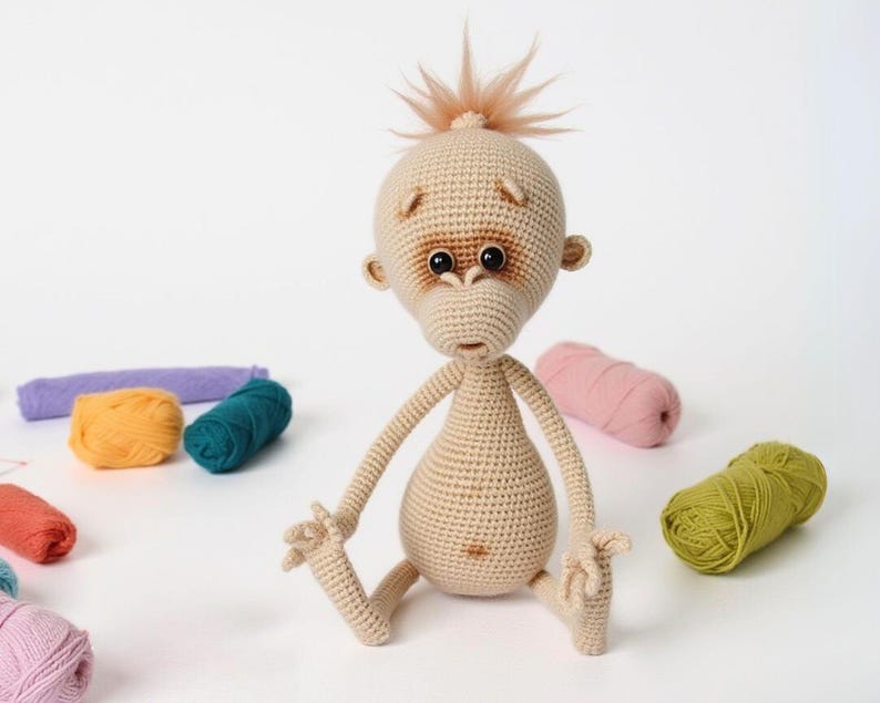 May include: A crocheted orangutan toy with brown eyes and a light brown body. The toy is sitting with its legs crossed and its arms outstretched. The toy is surrounded by balls of yarn in various colors.