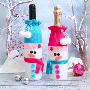 May include: Two snowman-themed bottle covers. One has a blue hat and scarf, the other a pink hat and blue scarf. Both have black button eyes, carrot noses, and red berry accents. The covers are white with blue and pink accents, and white pom-poms.