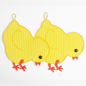 Op de afbeelding: Twee gehaakte gele kuiken ovenwanten met oranje voeten en zwarte ogen.