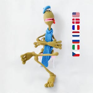 May include: A whimsical, handmade crochet creature in shades of olive green, blue, and pink. It wears a blue beret and scarf, with outstretched arms and legs. The creature has a pink mouth and tongue. A fun, unique decorative item.