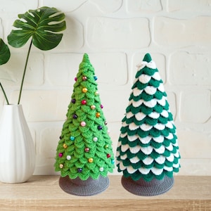 May include: Two crocheted Christmas trees, one green with colorful ornaments and one green and white, on a wooden surface.