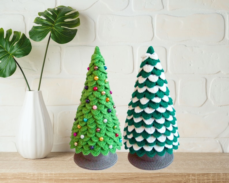 May include: Two crocheted Christmas trees, one green with colorful ornaments and one green and white, on a wooden surface.