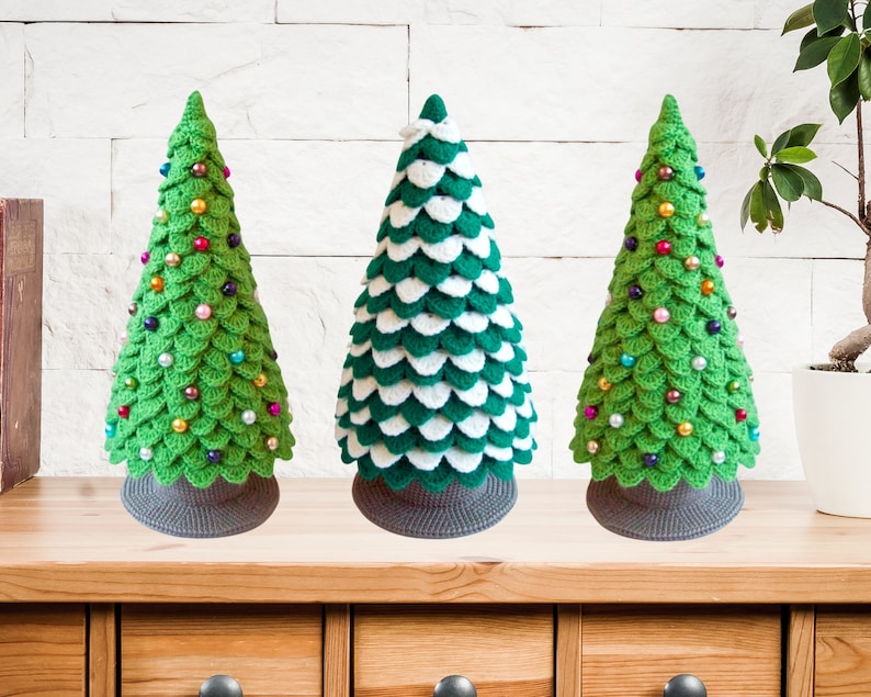 May include: Three crocheted Christmas trees, one green with colorful beads, one green and white with white beads, and one green with colorful beads. The trees are on a wooden shelf with drawers.