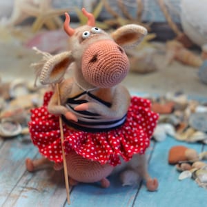 May include: A crocheted cow toy wearing a red and white polka dot skirt and a striped shirt. The cow has large eyes and a playful expression. The toy is sitting on a wooden surface with seashells scattered around it.