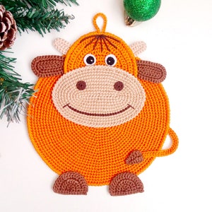 Op de afbeelding: Oranje gehaakte koe potdeksel met bruine oren en hoeven.