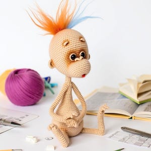 May include: A crocheted monkey toy with a brown face, orange and blue yarn hair, and a red mouth. The monkey is sitting on a white surface with yarn balls and books in the background.
